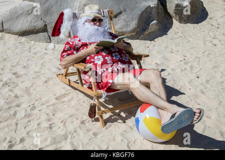 Weihnachtsmann in rot Badehose ans Hawaiian shirt am Sandstrand mit Stroh Hut und Sonnenbrille Buch lesen mit tropischen Drink. Stockfoto