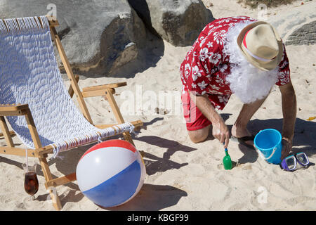 Weihnachtsmann in rot Badehose ans Hawaiian shirt am Sandstrand mit Stroh Hut und Sonnenbrille spielen mit Schaufel und blass. Stockfoto