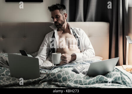 Gutaussehende kaukasische Männer, die im Bett mit mehreren Computern und mobilen Wireless-Geräten arbeiten, während sie eine Tasse Kaffee halten. Stockfoto