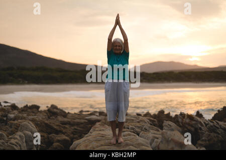 Ältere Frau Yoga am Meer Felsen bei Sonnenuntergang Stockfoto