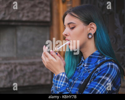 Junge Punk Mädchen rauchen Zigarette durch die holztür. Hipster mit blau gefärbten Haaren. Frau mit Piercing in der Nase, violett Linsen, Ohren Tunnel und ungewöhnliche Stockfoto