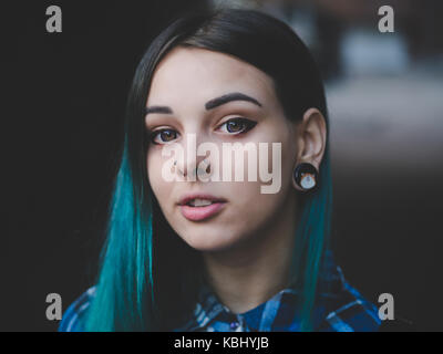 Street Punk oder Hippie Mädchen mit blau gefärbten Haaren. Frau mit Piercing in der Nase, violett Linsen, Ohren Tunnel und ungewöhnliche Frisur steht in der Stadt. Stockfoto