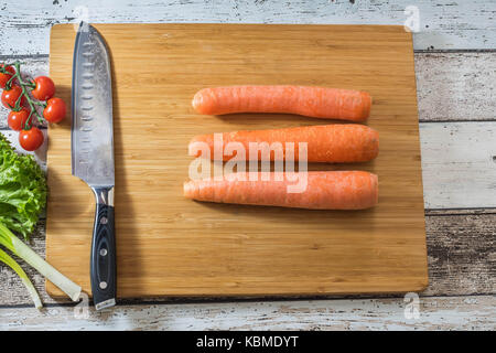 Santoku Küchenmesser auf einem Schneidebrett mit frischem Gemüse: Karotten, Tomaten, Salat und grüne Zwiebel auf einem hölzernen Hintergrund Draufsicht Stockfoto