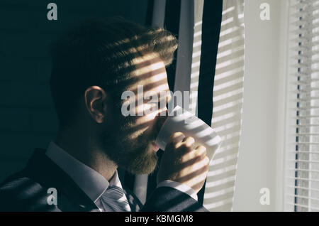 Hübscher junger Bartgeier Geschäftsmann ist, schauen aus dem Fenster und trinken Kaffee Stockfoto