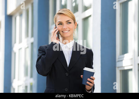 Lächelnden jungen Geschäftsfrau mit Handy beim Tragen verfügbare Kaffeetasse Stockfoto