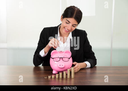 Junge Geschäftsfrau einfügen Münze in Sparschwein mit Münzen Stapel auf dem Schreibtisch Stockfoto