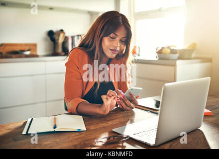 Lächelnden jungen afrikanischen weibliche Unternehmer und das Senden von SMS-Nachrichten, die an einem Notebook arbeitet, während an ihrem Küchentisch sitzen Stockfoto