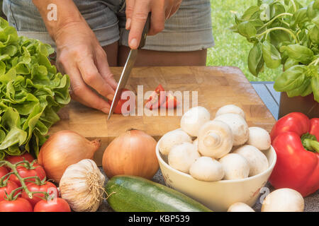 Vorderansicht des Schneiden eine Tomaten auf einem Holzbrett. Verschiedene Arten von Gemüse und Pilze liegen auf dem Tisch. Stockfoto