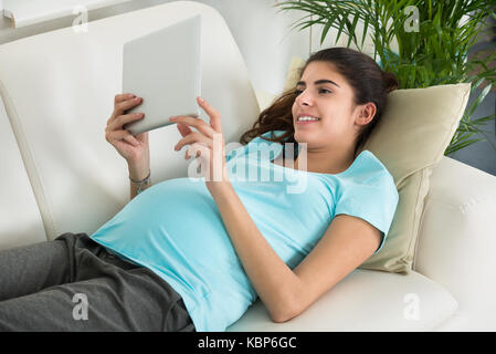 Gerne schwangere Frau mit digitalen tablet, während auf dem Sofa zu Hause liegen Stockfoto