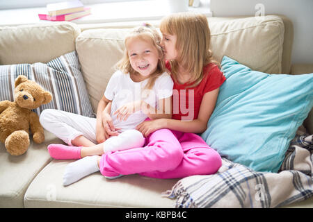 Porträt von zwei glücklichen kleinen Schwestern streicheln zärtlich auf bequeme Couch im Wohnzimmer zu Hause sitzen und Lächeln an Kamera Stockfoto