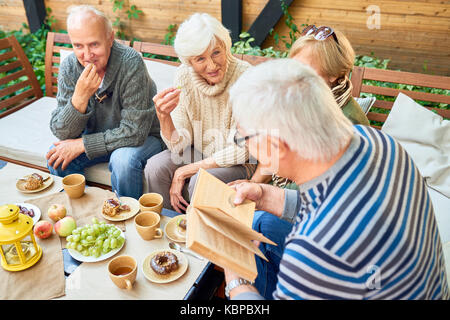 Gruppe von fröhlichen älteren Freunden Spaß im Freien: Sie genießen köstliche Kuchen und duftenden Kaffee, während ältere Mann im gestreiften Pullover lesen. Stockfoto