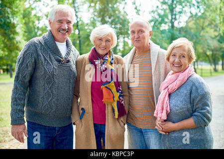 Group Portrait von fröhlichen älteren Freunde tragen warme Kleidung an Kamera suchen mit breitem Lächeln, während mit angenehmen Spaziergang am sonnigen Green Park Stockfoto