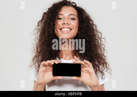 In der Nähe Bild von lächelnden curly Frau, leeren Bildschirm des Smartphones, und wenn man die Kamera über grauer Hintergrund Stockfoto