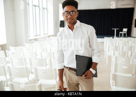 Bild von fröhlichen jungen schönen afrikanischen Mann in Office. Suchen Kamera holding Zwischenablage. Stockfoto