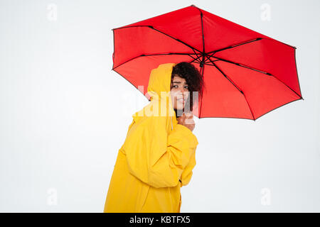 Junge afrikanische Frau in Regenmantel versteckt sich hinter Regenschirm, und wenn man die Kamera auf weißem Hintergrund Stockfoto
