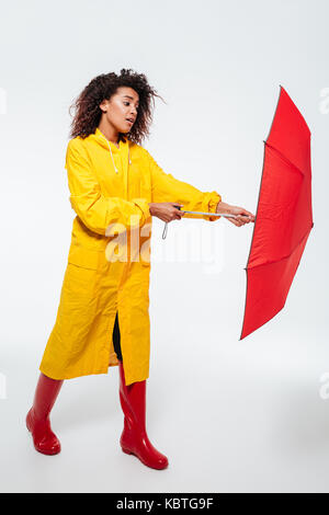 Volle Länge Bild der afrikanischen Frau verwirrt in Regenmantel Eröffnung Regenschirm auf weißem Hintergrund Stockfoto