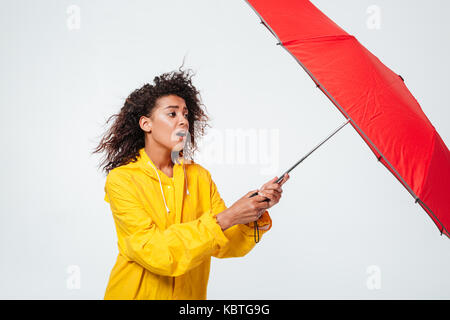 Seitenansicht des verwirrt afrikanische Frau in Regenmantel holding Regenschirm auf weißem Hintergrund Stockfoto