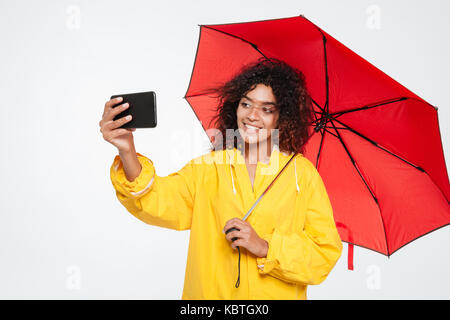 Gerne afrikanische Frau in Regenmantel versteckt unter Dach und selfie auf dem Smartphone auf weißem Hintergrund Stockfoto
