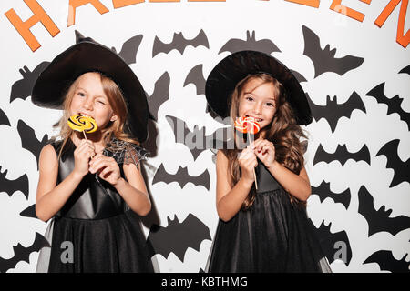 Paar Zwei lächelnde Mädchen in Halloween Kostüme essen Lutscher gekleidet, während mit Fledermäusen auf einem Hintergrund posiert Stockfoto
