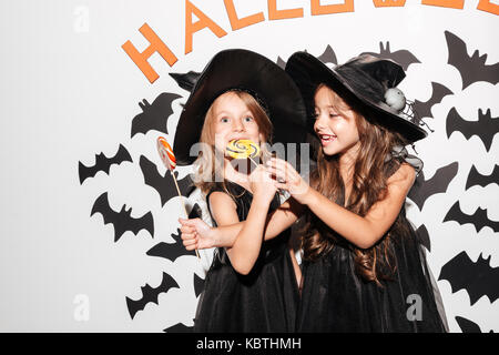 Paar von zwei entzückenden kleinen Mädchen in Halloween Kostüme essen Lutscher gekleidet, während mit Fledermäusen auf einem Hintergrund posiert Stockfoto