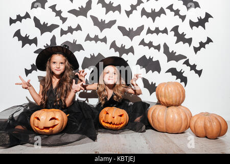 Paar von zwei kleinen Mädchen in Halloween Kostüme posiert während der Sitzung mit Kürbissen und Fledermäuse auf einem Hintergrund gekleidet Stockfoto