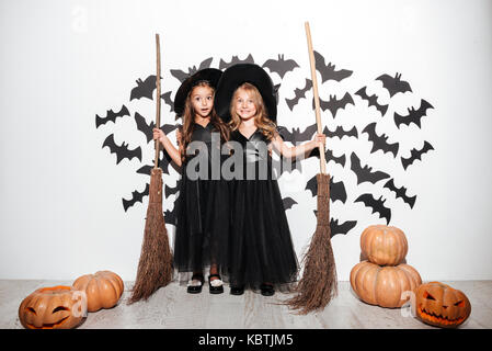 Paar zwei glückliche Lustige kleine Mädchen in Halloween Kostüme Besen halten und mit Fledermäusen und Kürbisse, die auf einem Hintergrund posiert gekleidet Stockfoto