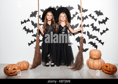 Paar zwei lustige kleine Mädchen in Halloween Kostüme Besen halten und mit Fledermäusen und Kürbisse, die auf einem Hintergrund posiert gekleidet Stockfoto