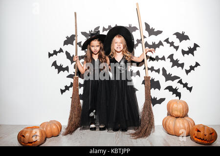 Paar von zwei kleinen Mädchen in Halloween Kostüme Besen halten und mit Fledermäusen und Kürbisse, die auf einem Hintergrund posiert gekleidet Stockfoto