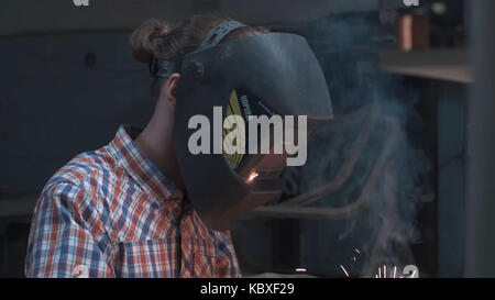 Junger Mann mit Schutzmaske Schweißen in einer Fabrik. Eine junge Brünette mann Schweißer auf ein schwarzes T-Shirt, in einem schweissverfahren Maske und Einbrecher, brüht einen Metall Schweißen Maschine in der Werkstatt Stockfoto