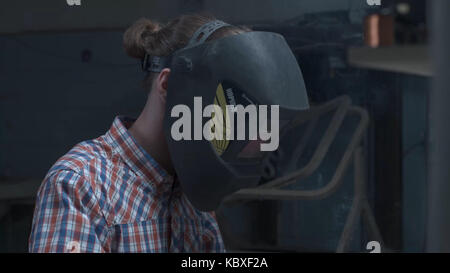 Junger Mann mit Schutzmaske Schweißen in einer Fabrik. Eine junge Brünette mann Schweißer auf ein schwarzes T-Shirt, in einem schweissverfahren Maske und Einbrecher, brüht einen Metall Schweißen Maschine in der Werkstatt Stockfoto