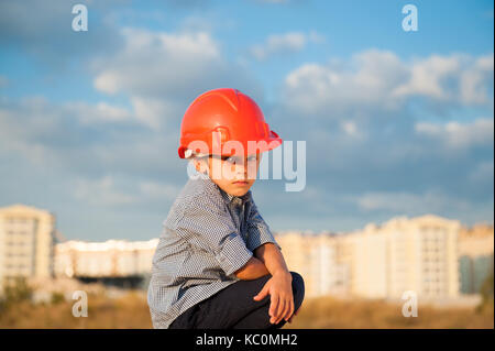 Cute Ernst kleiner Junge trägt orange Helm, der auf dem neuen Gebäude und schönen Himmel Hintergrund bei warmen Sonnenuntergang Stockfoto