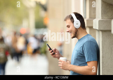 Profil von einer glücklichen Kerl hören Musik auf Linie mit Kopfhörern und einem smart phone lehnte sich an eine Wand auf der Straße Stockfoto