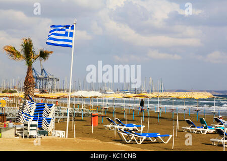 Strand in Larnaca Finikoudas mit Liegestühlen. Mit griechischer Flagge und Marina im Hintergrund. Stockfoto