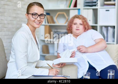 Porträt der schönen Psychologin Brille mit Klemmbrett in der Therapie Büro posing lächelnd in die Kamera, mit übergewichtigen Patienten in Backgr Stockfoto