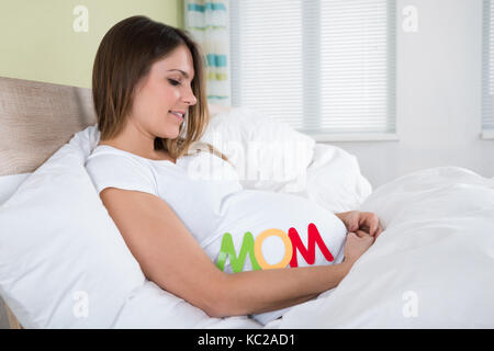 Junge schwangere Frau mit Text Mama im Bett sitzen Stockfoto