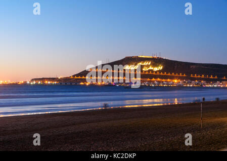 Blick auf langen, breiten Strand in Agadir City bei Nacht, Marokko. Der Berg trägt die Inschrift in Arabisch: Gott, Vaterland, König Stockfoto
