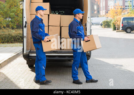 Lächelnden jungen Lieferung Männer, die Kartons während ausserhalb Fahrzeug zu Fuß Stockfoto