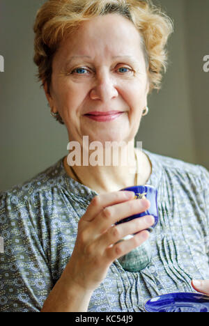 Attraktive Frau mittleren Alters Brille stends, lesen ein Buch in Ihrem Wohnzimmer Stockfoto