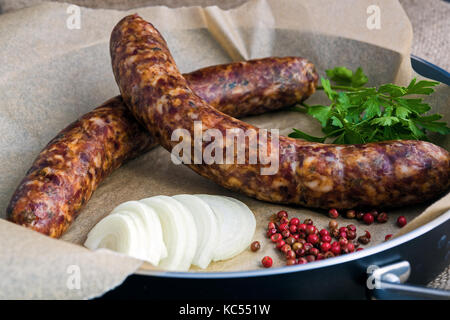 Zwei rohe Würste in einer Pfanne mit Zwiebeln, Gewürzen und Petersilie Stockfoto