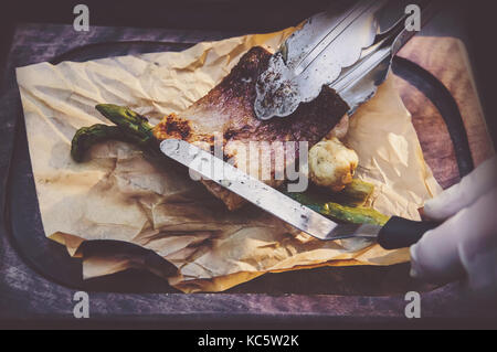 Prozess der Küche Fisch. Hände Köche in saubere, weiße Handschuhe setzt gegrillten Fisch auf dem Papier liegen auf dem Board. Stockfoto