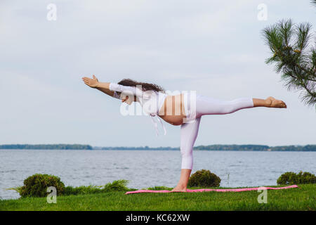 Schwangeren Yoga. Frauen, die verschiedenen Übungen im Park auf der Wiese, Atmung, Dehnung, Statik. Wald und Fluss auf Hintergrund. Konzept der Gesundheit Stockfoto