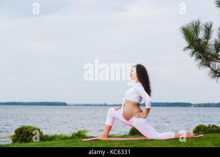 Schwangeren Yoga. Frauen, die verschiedenen Übungen im Park auf der Wiese, Atmung, Dehnung, Statik. Wald und Fluss auf Hintergrund. Konzept der Gesundheit Stockfoto