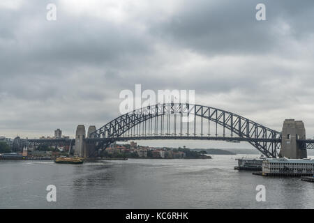 Sydney, Australien - 21. März 2017: Sydney Harbour Bridge unter grauen regnerischen Himmel. Dunkler grau Wasser vor mit der Fähre. Stockfoto