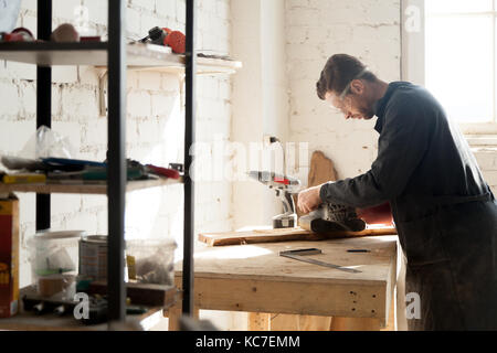 Seitenansicht junger ernster Schreinerschleifbretter, Mann in Arbeitskleidung und Schutzbrille, die Holz in seiner Werkstatt mit Geräten verarbeiten. Zimmerei Stockfoto
