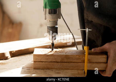 Nahaufnahme Foto von zwei Stücke von trockenem Holz befestigt Rohrschelle an der Werkbank und mit elektrischen Bohrmaschine gebohrt. Schreiner in einer Schreinerei arbeiten Werkstatt. Beruf Stockfoto