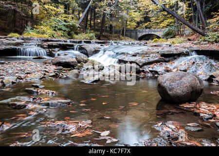 Anmutigen Wasserfällen Strömung um einen Stein Gehweg und gebogene Brücke an Wolf Creek, Letchworth State Park, NY Stockfoto