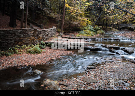 Anmutigen Wasserfällen Strömung um einen Stein Gehweg und gebogene Brücke an Wolf Creek, Letchworth State Park, NY Stockfoto