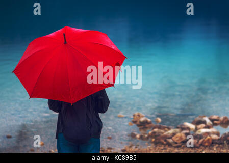 Frau mit roten Dach betrachtet auf Regen vor einem See. traurig und einsam weibliche Person in die Ferne schauen. Stockfoto