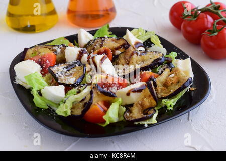 Auberginen Salat mit Tomaten, Mozzarella, Eisbergsalat mit Balsamico glace Essig und Olivenöl auf eine schwarze Platte auf einem weißen konkreten Hintergrund. Stockfoto