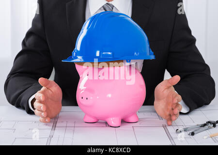 Nahaufnahme der männlichen Architekten schutz Sparschwein tragen Bau Helm auf Blueprint am Schreibtisch im Büro Stockfoto